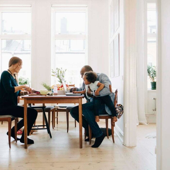 Äiti, isä ja taapero viettävät yhdessä vapaa aikaansa pöydän äärellä. Äiti tekee ruokaa ja isä selaa tablettia taapero sylissään Retta