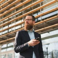 Mies kävelee lasisen toimistorakennuksen edessä ja selaa puhelinta Retta